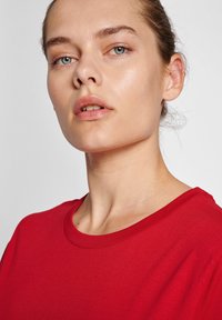 Rød bomulds t-shirt med rund halsudskæring og korte ærmer, vist på en model med lys hud og minimal makeup, mod en neutral baggrund.