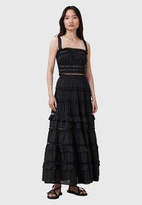 Robe longue noire à volants avec détails volantés, corsage smocké et accents en dentelle. Portée avec des sandales noires à lanières. Texture douce, design sans manches.