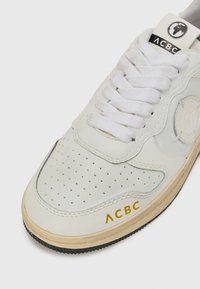 Sneaker bianco in pelle con punta perforata, lacci bianchi piatti, suola intermedia in crema testurizzata e suola esterna in gomma nera. Presenta il marchio dorato sul lato.