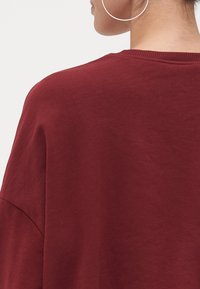 Suéter oversize burdeos con cuello redondo, costuras de hombros caídas y un tejido suave y texturizado. Diseño minimalista sin decoraciones.