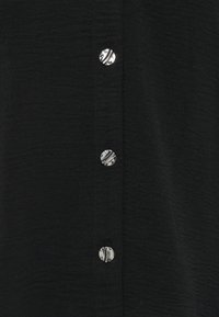 Μαύρο πουκάμισο με υφή, που διαθέτει μια κατακόρυφη σειρά γυαλιστερών ασημένιων κουμπιών με ριγέ μοτίβο. Το ύφασμα φαίνεται ελαφρύ και απαλό.