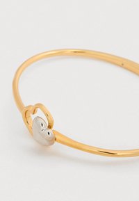 Βραχιόλι χειροπέδα σε χρυσή απόχρωση με λείο στρογγυλό σχήμα, που διαθέτει ασημένια καρδιά ως κούμπωμα και λεπτή, γυαλιστερή υφή.