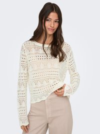 Giovane donna con capelli lunghi e scuri che indossa un top bianco all'uncinetto a maniche lunghe e pantaloni beige chiaro, tenendo l'orlo del top con una mano.