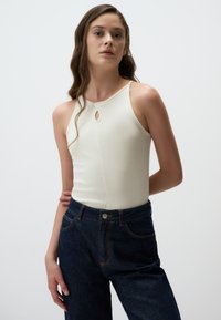 Top bianco senza maniche con dettaglio a chiave, abbinato a jeans in denim blu scuro a vita alta con cuciture a contrasto e chiusura frontale con bottone.