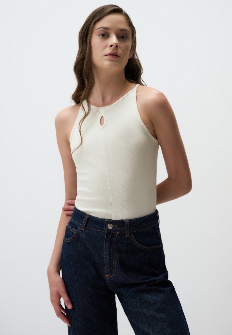 Top bianco senza maniche con dettaglio a chiave, abbinato a jeans in denim blu scuro a vita alta con cuciture a contrasto e chiusura frontale con bottone.