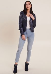 Giacca da biker in pelle blu navy con zip argentata, maglietta bianca a V, jeans skinny azzurri chiari e stivali alla caviglia blu navy con bordo decorativo.