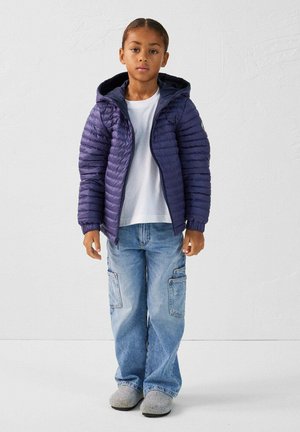 Veste doudoune violette avec une capuche, fermeture éclair, poignets côtelés, portée sur un t-shirt blanc, associée à un jean cargo bleu clair et des chaussons gris.