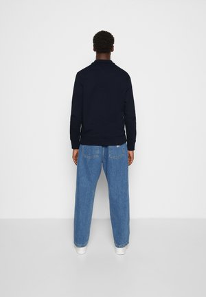 Mann steht vor einer weißen Wand, trägt eine schwarze Jacke, lockere blaue Jeans und weiße Turnschuhe, die Hände ruhen an den Seiten.