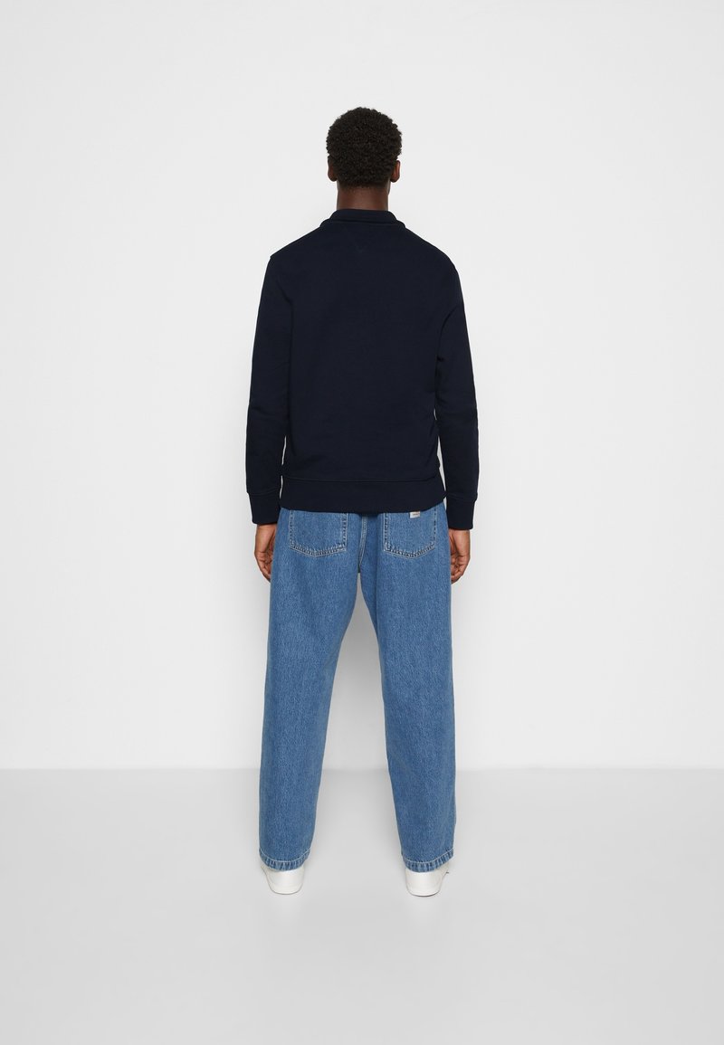 Homme debout face à un mur blanc, portant une veste noire, un jean bleu ample et des baskets blanches, les mains reposant le long du corps.