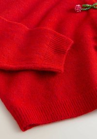 Roter Wollpullover mit einem gerippten Halsausschnitt und Bündchen. Verfügt über ein kleines dekoratives Ornament an der Schulter für zusätzliche Details. Weiche Textur.