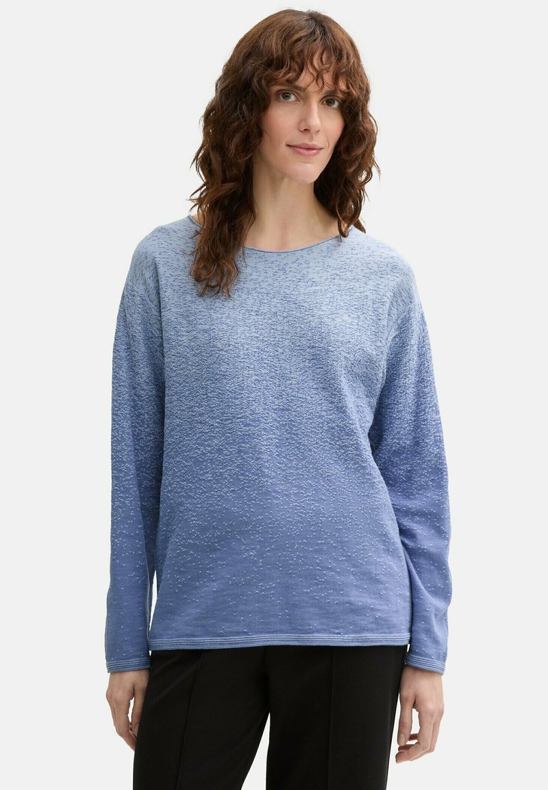 Hellblaues langärmliges Oberteil mit strukturiertem Muster, rundem Halsausschnitt und weichem Stoff. Darunter eine lockere schwarze Hose.