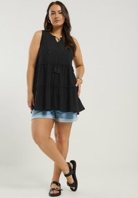 Black sleeveless top with a tiered design, featuring a drawstring neck and decorative tassels. Paired with light denim shorts and black sandals.
