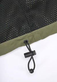 Tela de malla negra con pequeños agujeros, acentuada con un ribete inferior verde oliva. Cuenta con un cordón negro con un tope para ajuste.