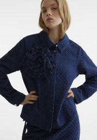 Giacca blu con texture, caratterizzata da una grande rosa in tessuto sul lato sinistro, con colletto e chiusura a bottoni.