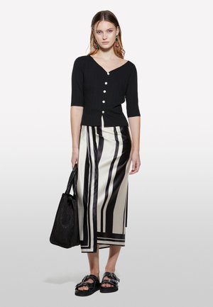 Schwarzes geripptes Oberteil mit kurzen Ärmeln und Knopfdetails, kombiniert mit einem langen gestreiften Rock in Beige und Schwarz. Das Model hält eine schwarze Tote-Tasche und trägt klobige Sandalen.
