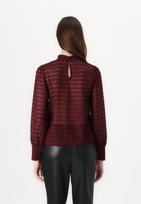 Bordeaux blonderbluse med lange ærmer, struktureret diamantmønster, nøglehulslukning bagpå og en tætsiddende talje. Sammen med sorte læderbukser.