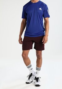 Kort blå idrottströja med vit logotyp, kombinerad med maroonshorts med ett strukturerat mönster. Bärs med randiga vita strumpor och svarta skor.