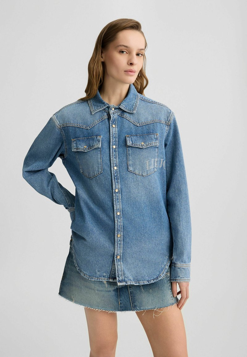 Camicia in denim di colore azzurro chiaro con colletto classico, chiusura anteriore con bottoni, due tasche sul petto e dettagli ricamati sulla tasca sinistra.