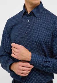 Camicia button-up blu navy con un piccolo motivo a pois bianchi, colletto, maniche lunghe e bottoni sui polsini e sulla patta frontale.