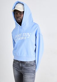 Ljust blå kort hoodie med vit "tommy jeans" logotyp, med dragsko i huvan och ribbad nederkant, kombinerad med åtsittande grå jeans.