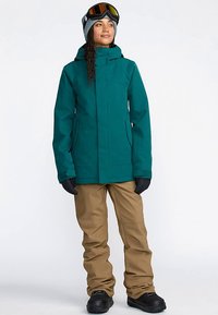 A teal waterproof jacket with a hood, paired with tan snow pants and black gloves. The outfit is completed with ski goggles on the forehead.