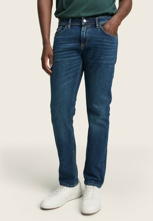 Jeans de mezclilla azul con un corte recto, que cuentan con cinco bolsillos, un ligero desgastado y acentos de costura superior. Combinados con zapatillas blancas.