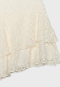 Tissu en dentelle couleur crème avec motif floral, présentant des bords superposés festonnés et une texture délicate et semi-transparente.
