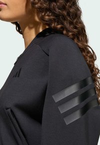 Žena u crnoj jakni na zip s tri crne pruge na rukavu i suptilnim logotipom na prsima, kovrčava kosa vidljiva sa strane.