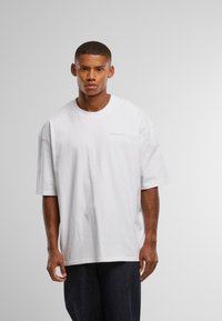 Bílé volné bavlněné tričko s kulatým výstřihem a krátkými rukávy. Na hrudi má jemné a nenápadné logo. Nošeno s tmavými džínovými kalhotami.