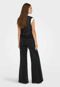 Cappotto in maglia nera con una vestibilità ampia, caratterizzato da un design perforato, indossato sopra una camicia bianca; abbinato a jeans neri a gamba larga.