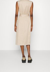 Beżowa sukienka bez rękawów z paskiem w talii, do kolan, wykonana z gładkiej tkaniny. Do tego jasnobrązowe buty z grubą czarną podeszwą.