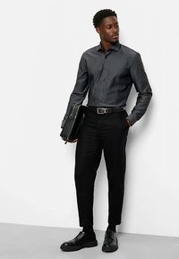 Dunkelgraues Hemd mit Knöpfen, schwarze schmal geschnittene Hose, schwarze Lederschuhe, hält eine schwarze Aktentasche, vor schlichtem Hintergrund.