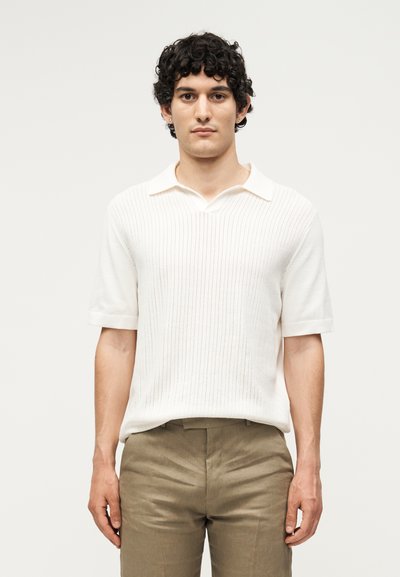Jeune homme aux cheveux bouclés et foncés, portant un polo en maille blanc à manches courtes et un pantalon marron clair, debout devant un fond clair uni.