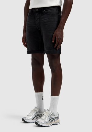 Man wearing black rolled denim shorts, white high socks, and silver running shoes standing against a plain background.