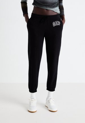 Osoba ubrana w czarne spodnie dresowe GAP z elastycznymi ściągaczami, białe skarpetki i białe trampki, stojąca z rękami w kieszeniach.