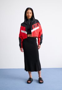 Czerwona i czarna sportowa kurtka zapinana na zamek o krótkim kroju z białymi akcentami, czarny crop top, czarna spódnica midi i czarne klapki na niebieskiej podłodze.