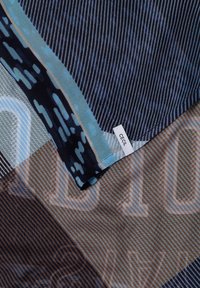 Gros plan d’un tissu plié avec des motifs bleus et marron et une petite étiquette blanche portant la mention "CECIL" cousue au bord.