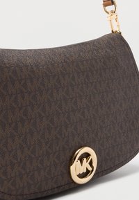 Sac à main en tissu marron avec un motif de logo subtil, forme incurvée et un accent de logo circulaire en ton or. Dispose d'une bandoulière amovible.