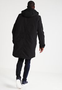 Mann, der weggeht, gekleidet in eine schwarze kapuzenparka, dunkle Hose und marineblaue Sneakers mit weißen Sohlen vor einem schlichten weißen Hintergrund.