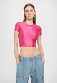 Rožinė satino trumpa palaidinė su trumpomis rankovėmis, turinti logotipo spaudą centre, derinama su šviesiai mėlynomis džinsinėmis kargo kelnėmis su mygtukų detalėmis.