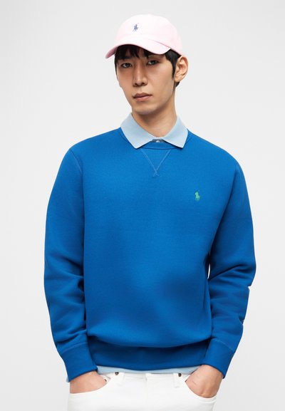 Άνδρας που φοράει έντονο μπλε φούτερ πάνω από ένα ανοιχτό μπλε πουκάμισο με γιακά, με λευκό παντελόνι και ανοιχτό ροζ καπέλο, στέκεται μπροστά σε λευκό φόντο.