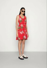 Vestido vermelho sem mangas com um padrão floral, cintura ajustada e saia rodada. Combinado com sandálias brancas com sola de plataforma preta.