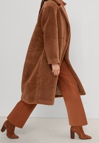 Abrigo marrón, esponjoso y de gran tamaño con cuello tipo chal, combinado con pantalones anchos marrones y botas de tacón a juego, mostrado en una pose de caminar.