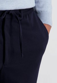 Close-up van een persoon die donkerblauwe joggingsbroek draagt, met hun rechterhand in de zak en een lichtblauwe mouwen van een trui zichtbaar.