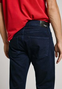 Dark denim jeans with a straight-fit design, featuring back pockets, contrast stitching, and a black leather patch on the waistband.