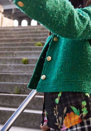 Woman wearing green textured coat and floral skirt holding metal railing while ascending outdoor stone stairs.