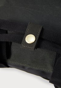 Close-up of black fabric strap with stitched loop and gold button engraved with "BARBOUR" on a dark textile background.