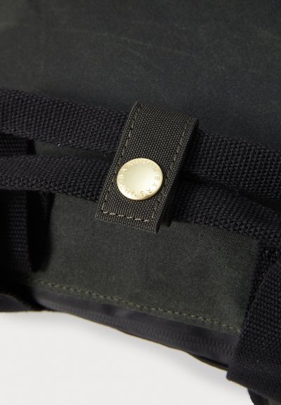 Gros plan sur une sangle en tissu noir avec une boucle cousue et un bouton doré gravé de "BARBOUR" sur un fond textile sombre.