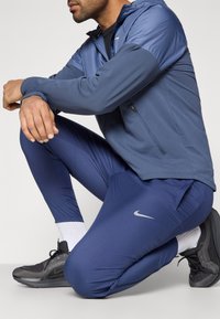 Άνδρας γονατιστός φορώντας μπλε αθλητική ζακέτα και παντελόνι, λευκές κάλτσες και μαύρα αθλητικά παπούτσια με κορδόνια.