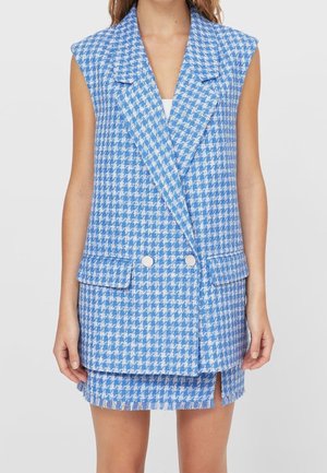 Femme portant un blazer sans manches à motif pied-de-poule bleu et blanc avec des boutons argentés et une mini-jupe assortie fendue à l'avant.